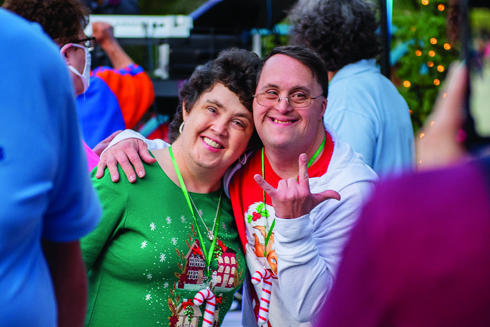 Two smiling adults pose together at The Arc Tampa Bay Foundation's annual Festival of Trees event in Clearwater, Florida. One person wears a festive green shirt with gingerbread houses and candy canes, while the other wears a white hoodie with holiday graphics and makes a playful hand gesture. Colorful lights and attendees can be seen in the background, creating a joyful, celebratory atmosphere.