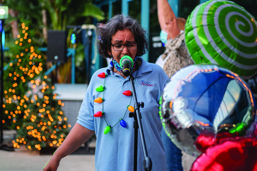 A woman sings into a microphone at The Arc Tampa Bay Foundation's annual Festival of Trees event in Clearwater, Florida. She wears a light blue Plato Academy shirt and a festive string of oversized holiday lights around her neck. Behind her are colorful balloons, a decorated Christmas tree with twinkling lights, and blurred attendees enjoying the celebration.