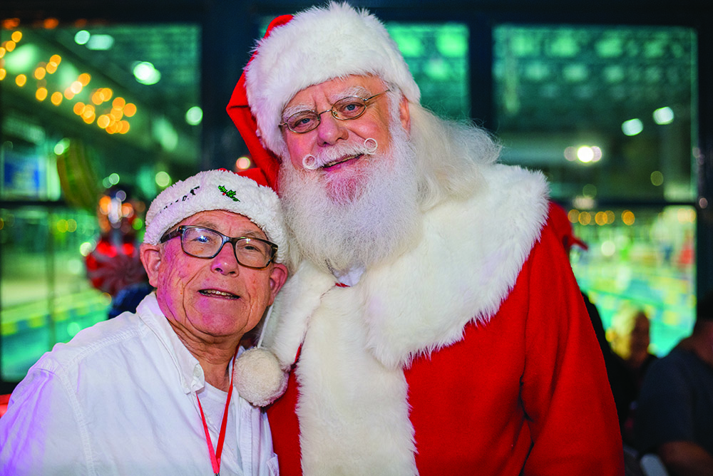 A man wearing a festive white shirt and Santa hat poses with Santa Claus at The Arc Tampa Bay Foundation's annual Festival of Trees event in Clearwater, Florida. Santa is dressed in his classic red and white suit with glasses and a cheerful smile. The background is filled with colorful lights and holiday decorations, creating a warm and joyful atmosphere.