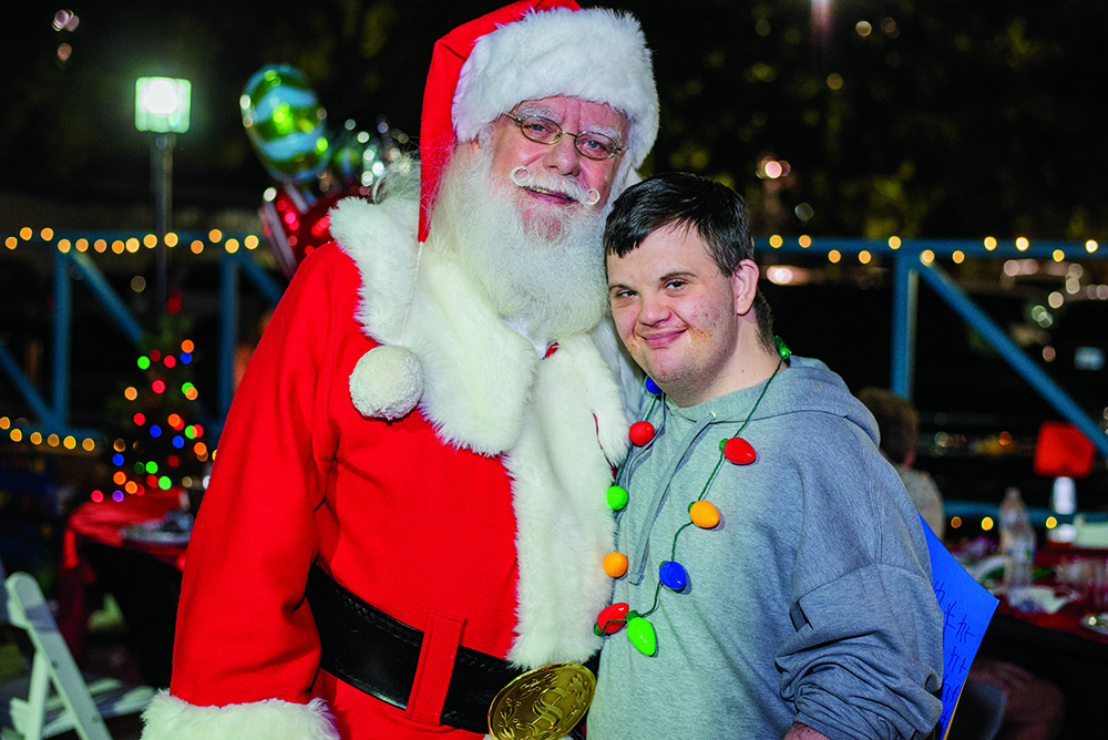 Santa Claus poses with a smiling young man wearing a gray hoodie and a colorful holiday light necklace at The Arc Tampa Bay Foundation's annual Festival of Trees event in Clearwater, Florida. The festive nighttime setting features twinkling string lights, decorated tables, and a glowing Christmas tree in the background, creating a joyful holiday atmosphere.