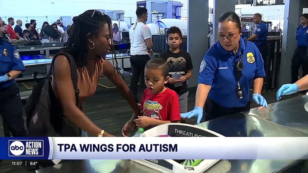 A mother helps her young son place items in a security bin at Tampa International Airport during The Arc Tampa Bay Foundation's Wings for Autism event. A TSA officer assists nearby, while other participants wait in line in the background. The event provides families with a practice run of the airport experience to help ease the stress of air travel for individuals with autism.