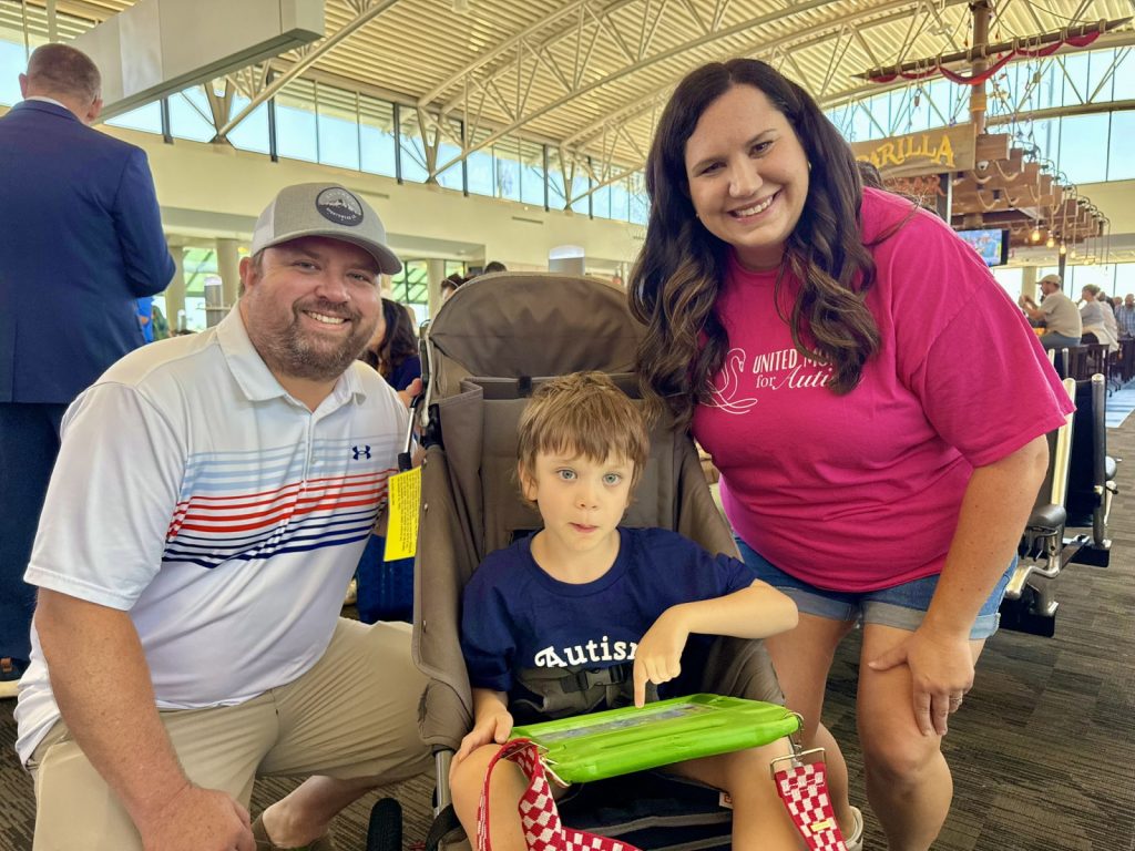 A smiling man and woman pose with a young boy seated in a stroller at Tampa International Airport during The Arc Tampa Bay Foundation's Wings for Autism event. The boy is wearing a navy blue shirt that says “autism” and is using a green tablet. The woman is wearing a bright pink shirt with the words “United for Autism.” The event helps families practice the airport experience in a supportive environment.