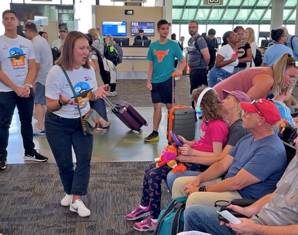 A volunteer wearing a Wings for Autism t-shirt engages with families seated at a gate inside Tampa International Airport during The Arc Tampa Bay Foundation's Wings for Autism event. A young girl wearing light-up headphones holds a tablet and sits beside adults, while travelers and volunteers move through the busy terminal in the background. The event provides a simulated travel experience to help individuals with autism become more comfortable with flying.