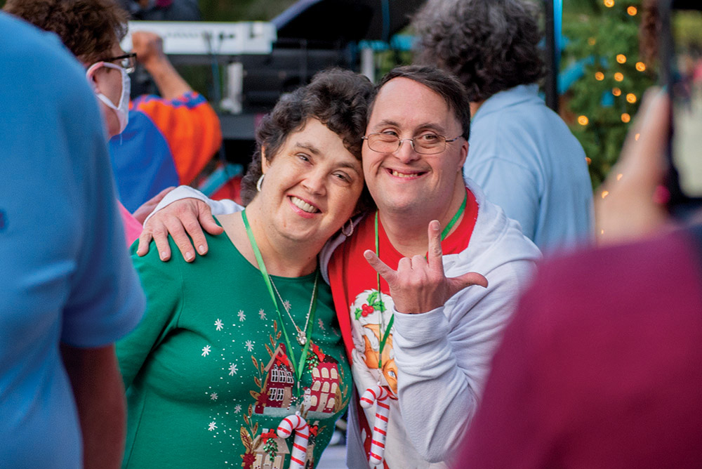 Two smiling adults pose together at The Arc Tampa Bay Foundation's annual Festival of Trees event in Clearwater, Florida. One person wears a festive green shirt with gingerbread houses and candy canes, while the other wears a white hoodie with holiday graphics and makes a playful hand gesture. Colorful lights and attendees can be seen in the background, creating a joyful, celebratory atmosphere.