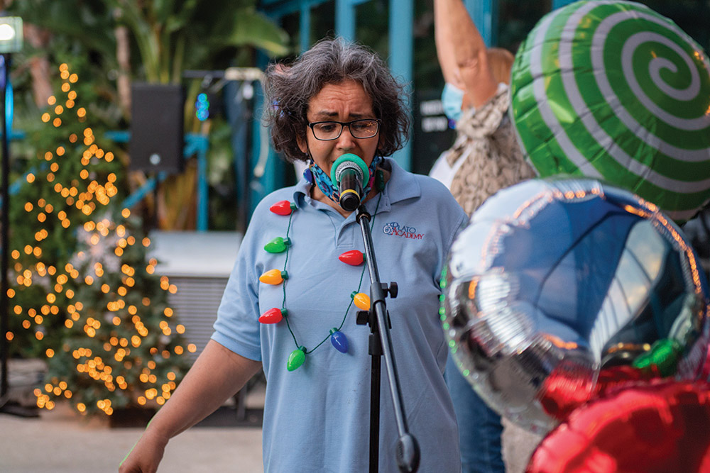 A woman sings into a microphone at The Arc Tampa Bay Foundation's annual Festival of Trees event in Clearwater, Florida. She wears a light blue Plato Academy shirt and a festive string of oversized holiday lights around her neck. Behind her are colorful balloons, a decorated Christmas tree with twinkling lights, and blurred attendees enjoying the celebration.