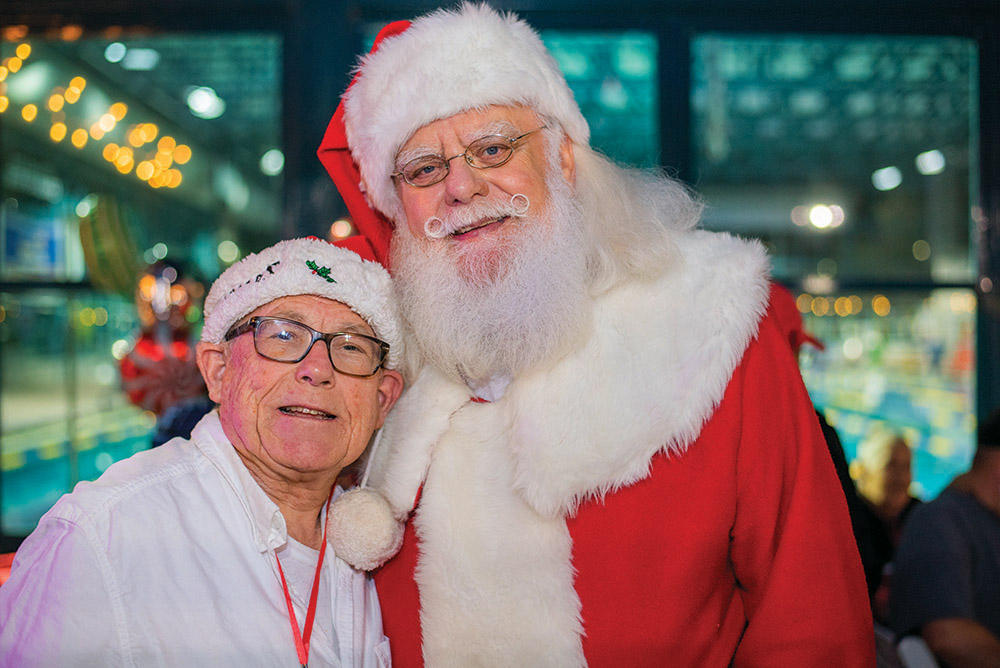 A man wearing a festive white shirt and Santa hat poses with Santa Claus at The Arc Tampa Bay Foundation's annual Festival of Trees event in Clearwater, Florida. Santa is dressed in his classic red and white suit with glasses and a cheerful smile. The background is filled with colorful lights and holiday decorations, creating a warm and joyful atmosphere.