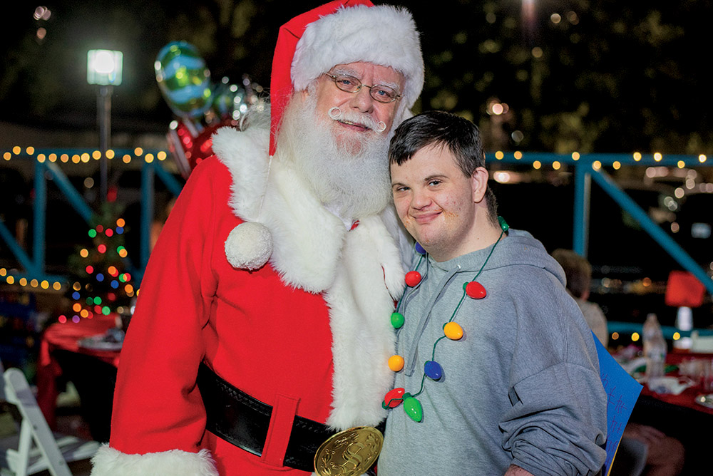 Santa Claus poses with a smiling young man wearing a gray hoodie and a colorful holiday light necklace at The Arc Tampa Bay Foundation's annual Festival of Trees event in Clearwater, Florida. The festive nighttime setting features twinkling string lights, decorated tables, and a glowing Christmas tree in the background, creating a joyful holiday atmosphere.